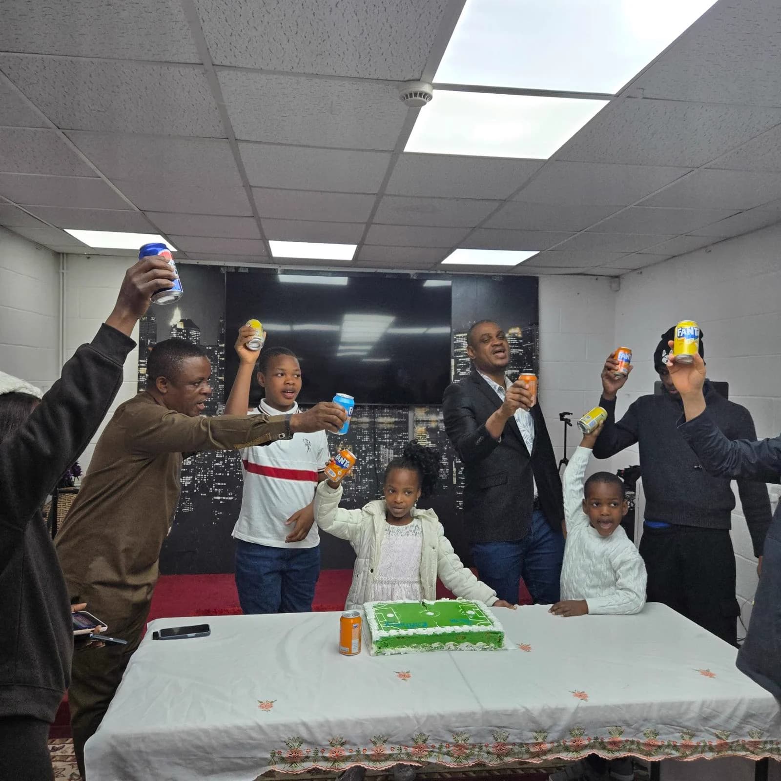 Members and children raising cans in a toast around a table with a green cake, in the worship hall.