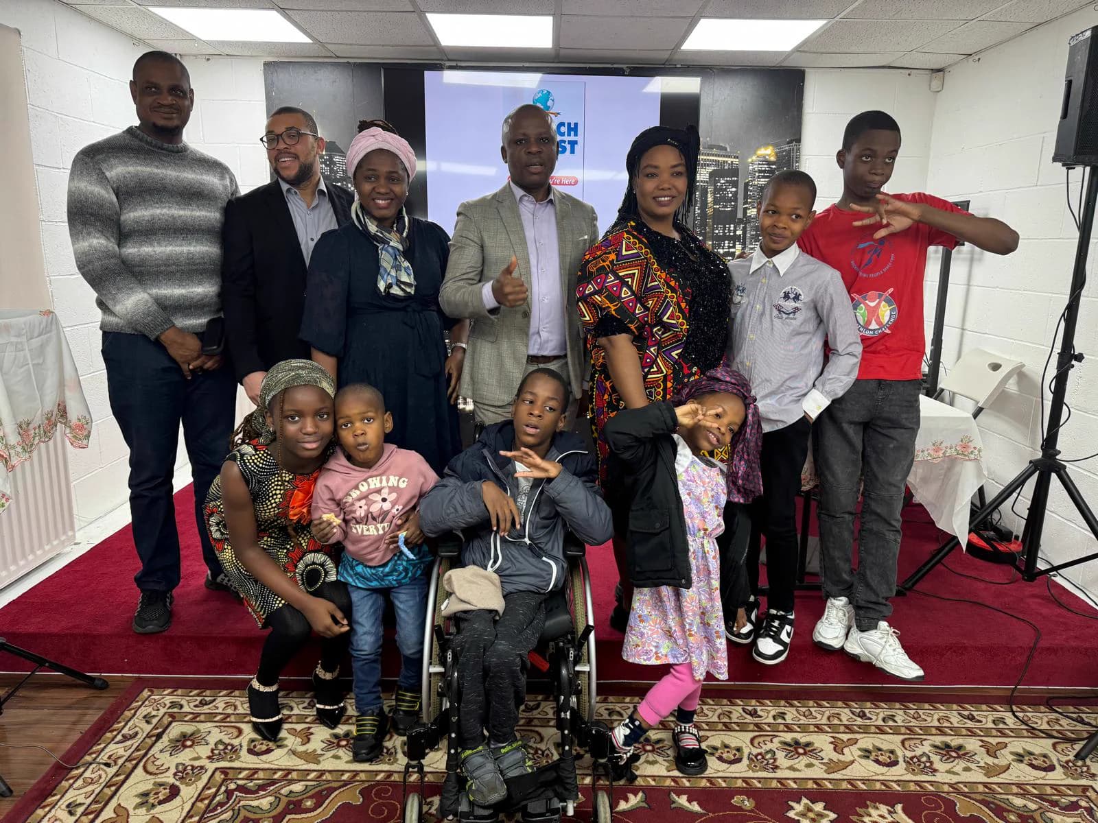 Members of all ages together on the red carpet of the worship hall, the church's emblem on the screen behind them.