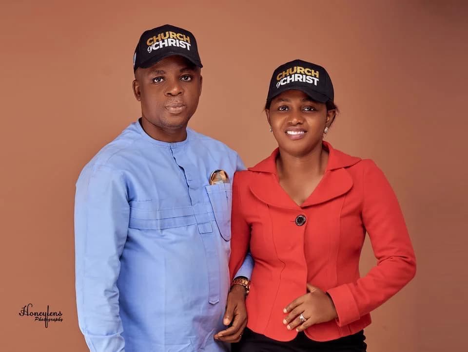 Kufre Okon in a pale blue outfit and a woman in a red jacket, both wearing black Church of Christ caps — a studio portrait.