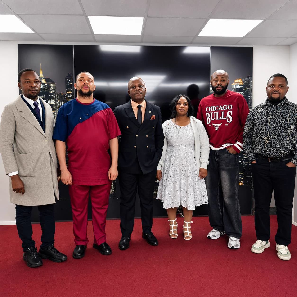 Six members standing together on the red carpet of the worship hall, in front of the night-skyline mural.