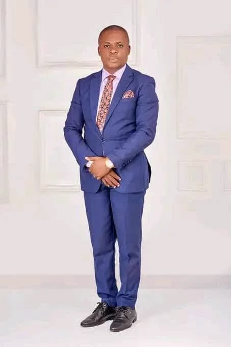 Kufre Okon standing full length in a blue suit and patterned tie, hands clasped.