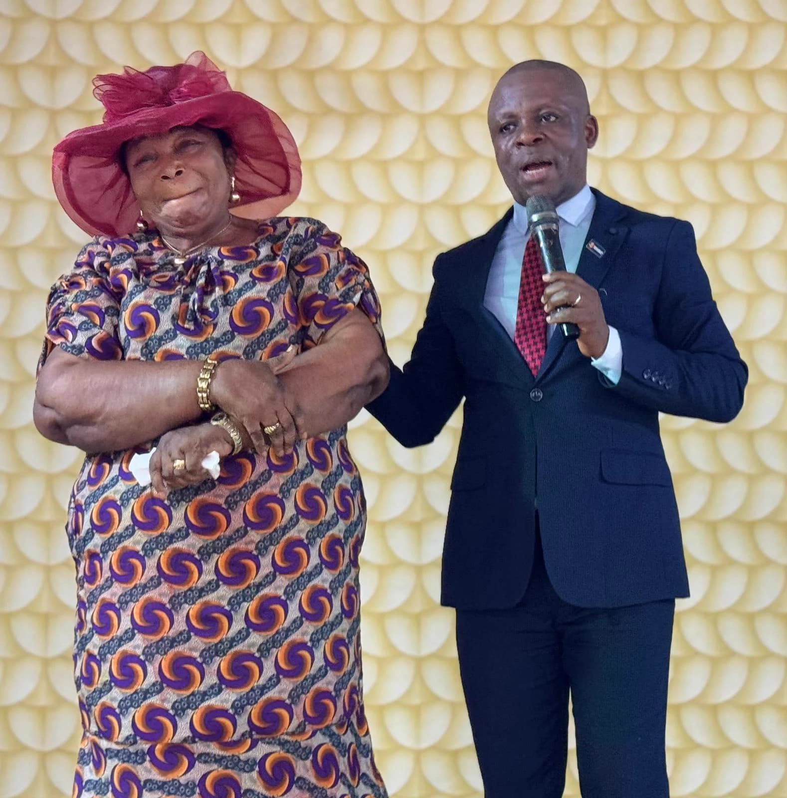 Kufre Okon in a dark suit speaking into a microphone beside a woman in a wide-brimmed maroon hat, at a community event.