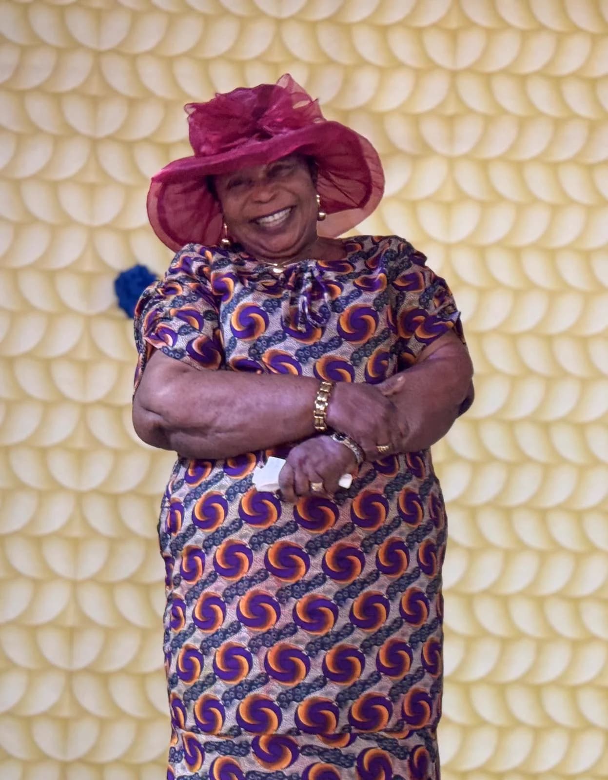 A woman in a wide-brimmed maroon hat and a patterned dress, arms folded, smiling.