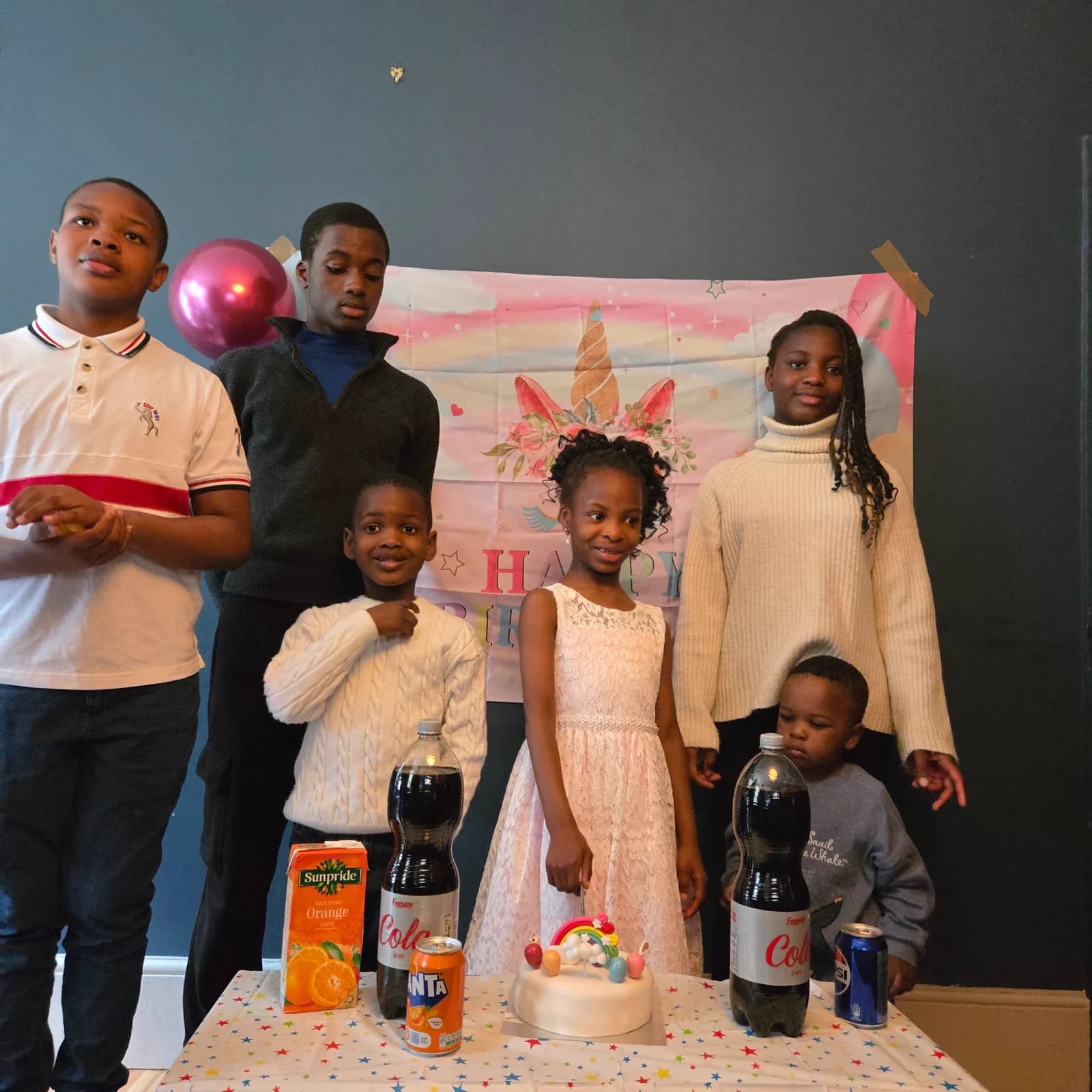 Children standing together at a birthday table in the hall, with a cake, drinks and a Happy Birthday banner behind them.