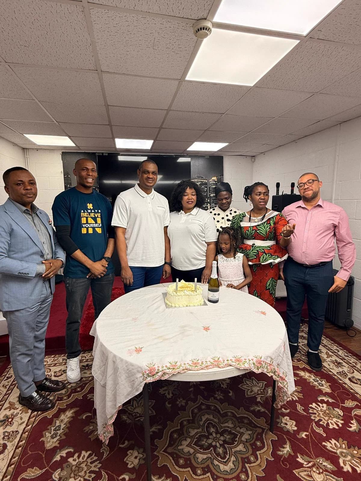 Members gathered around a table laid with a white cloth and a cake in the worship hall, a celebration after the gathering.