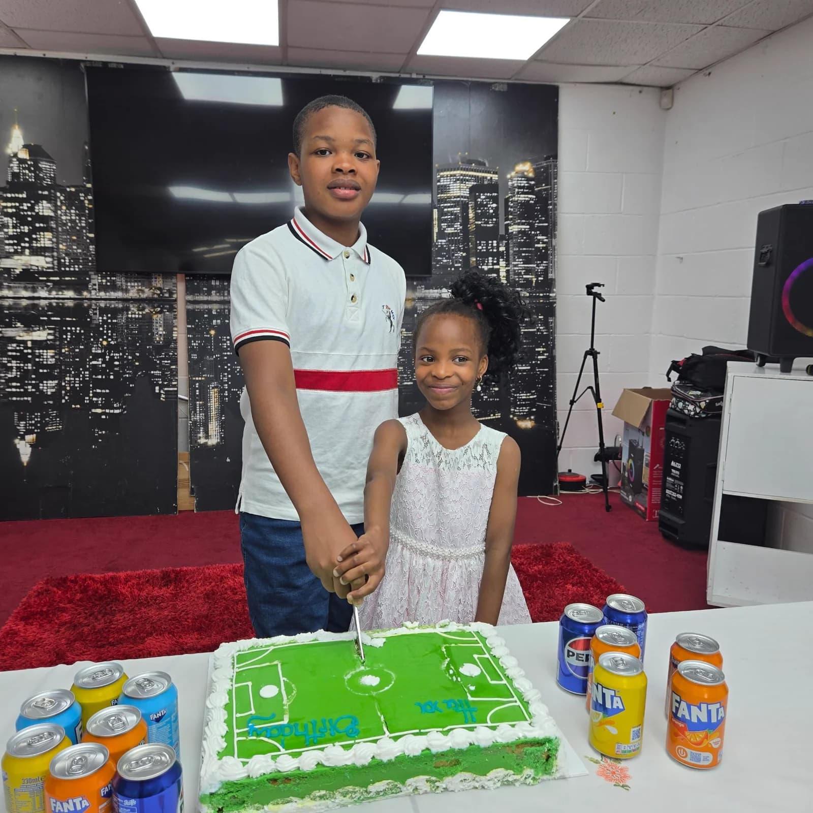 A boy and a girl cutting a green football-pitch birthday cake at a table in the worship hall.