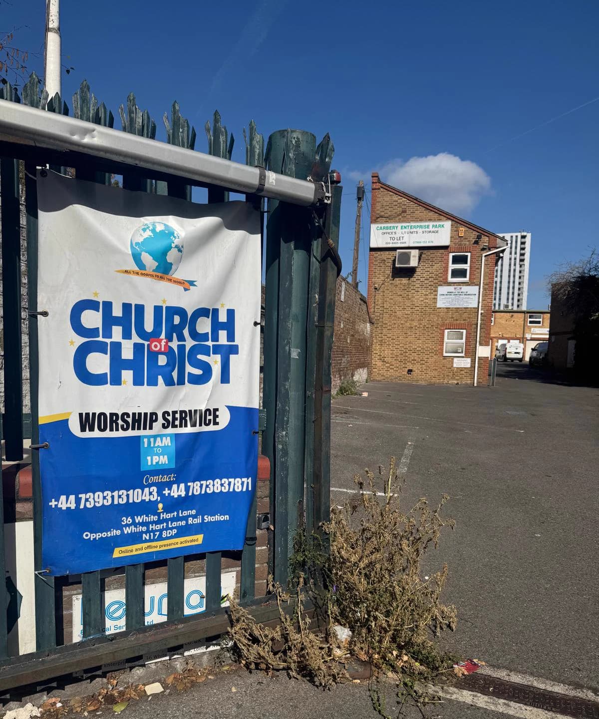 The entrance from the gate: the church's blue and white banner fixed to green railings, announcing the Sunday worship service, with the building behind.