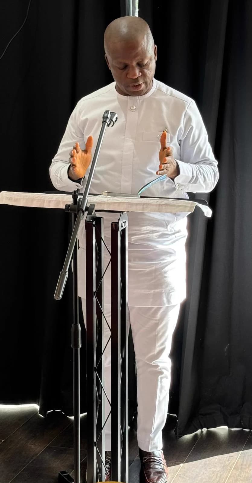 Kufre Okon in white at a lectern, reading from an open book into a microphone against a black curtain.