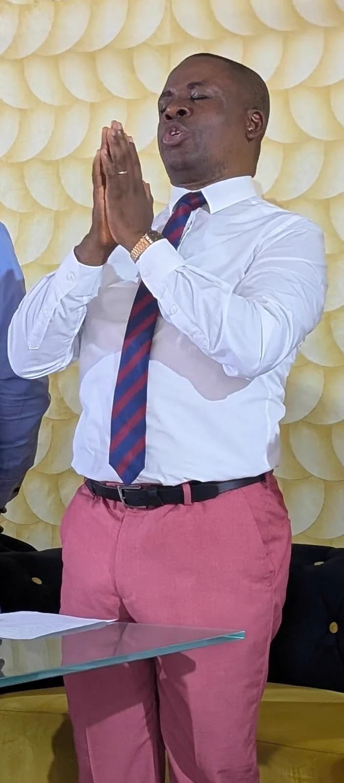 Kufre Okon in prayer, hands pressed together, in a white shirt and a rose-pink suit.