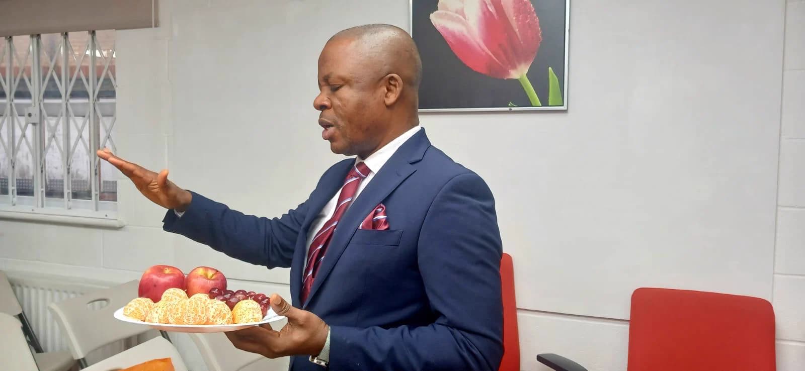 Kufre Okon teaching in the worship hall, in a blue suit and red tie, one hand raised as he speaks.