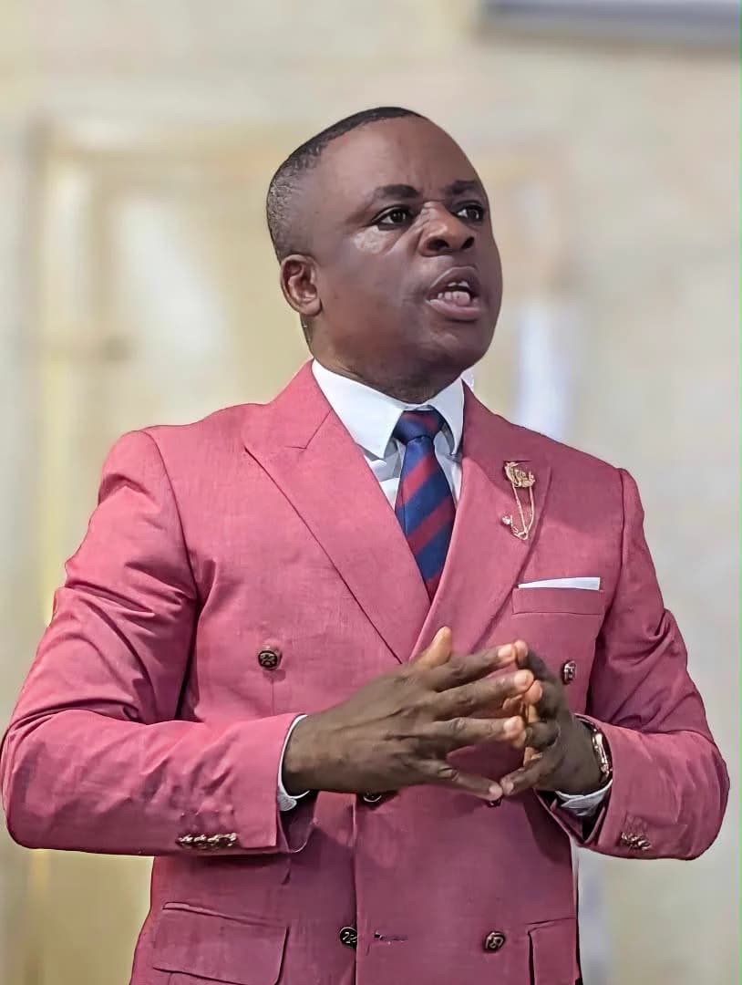 Kufre Okon, mid-sentence with his hands clasped, in a rose-pink double-breasted suit and a striped tie.