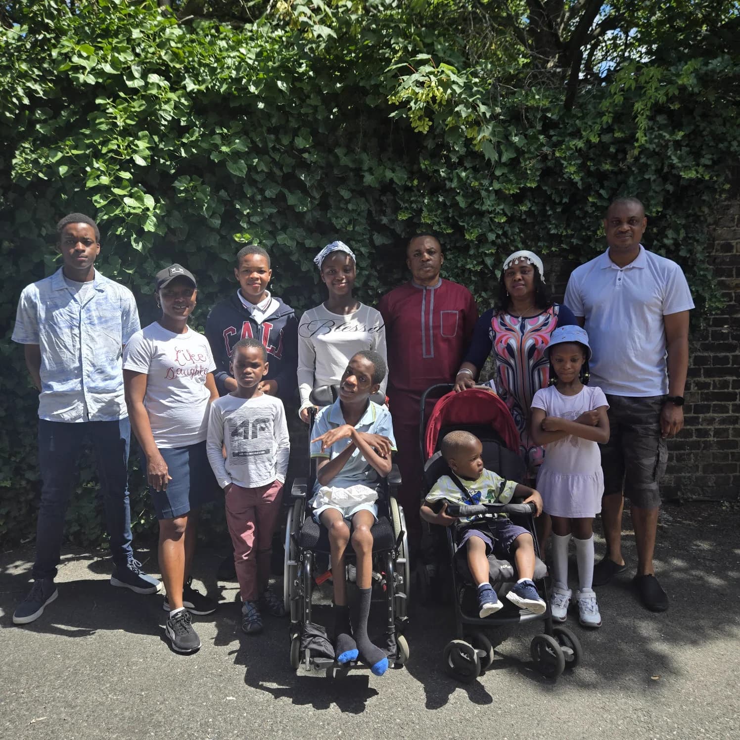 Members of all ages standing together outdoors in the sunshine against a wall of ivy, a child in a wheelchair and a baby in a pushchair at the front.