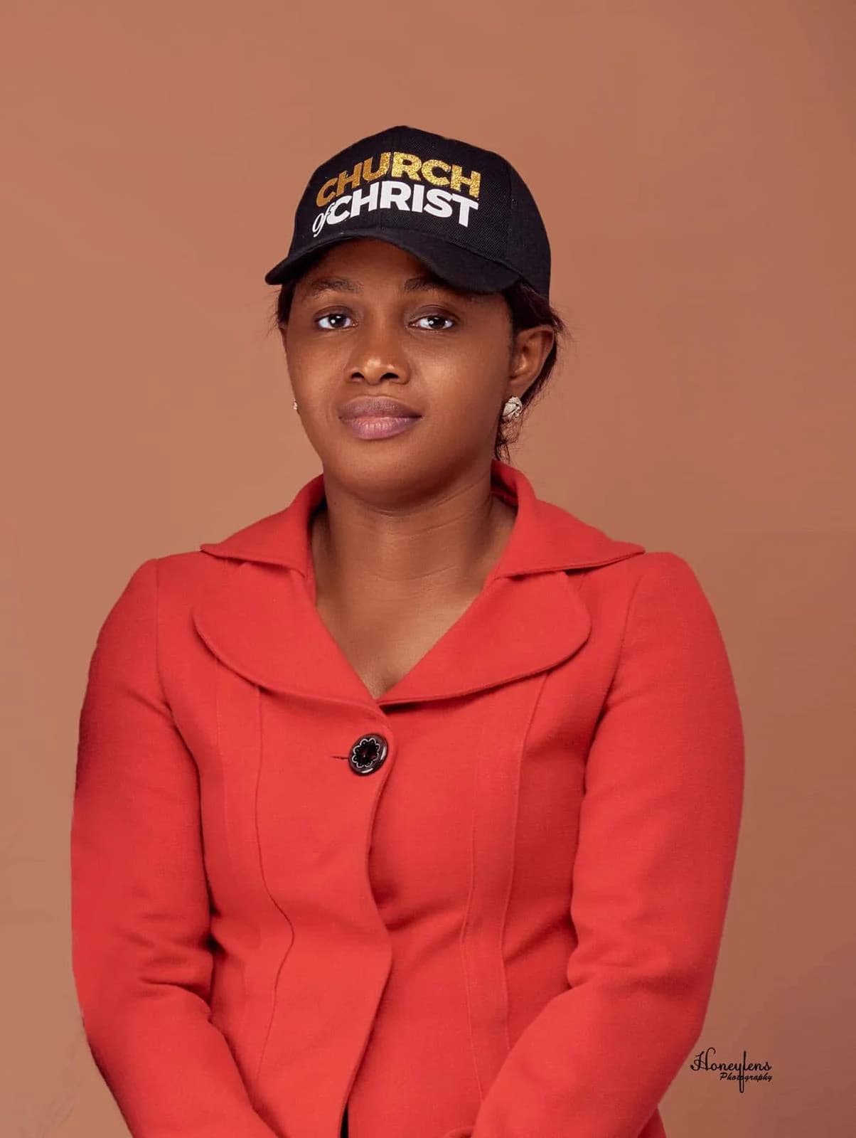 A woman in a red jacket and a black Church of Christ cap, hands folded, with a gentle smile — a studio portrait on a terracotta backdrop.