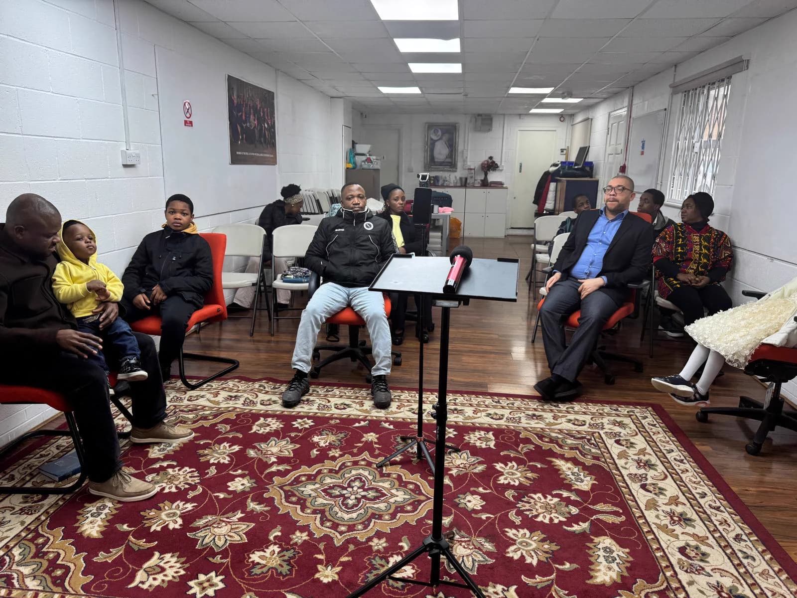 The congregation seated in the worship hall during a gathering, the lectern in the middle of the room and the red patterned carpet in the foreground.