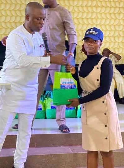 Kufre Okon, microphone in hand, handing a green bag of supplies to a young woman in a dark cap, in front of a patterned wall.