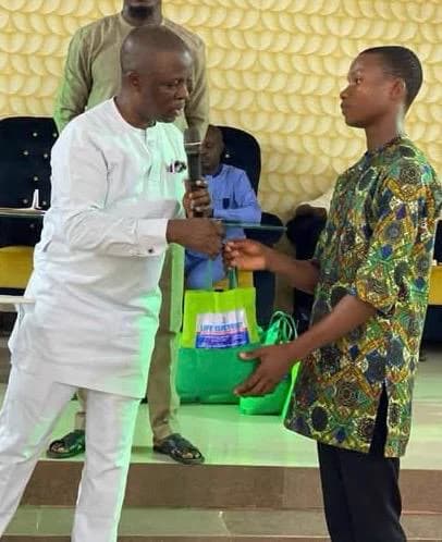 Kufre Okon, microphone in hand and dressed in white, handing a green bag of supplies to a young man in a patterned shirt, with more bags lined up behind them.
